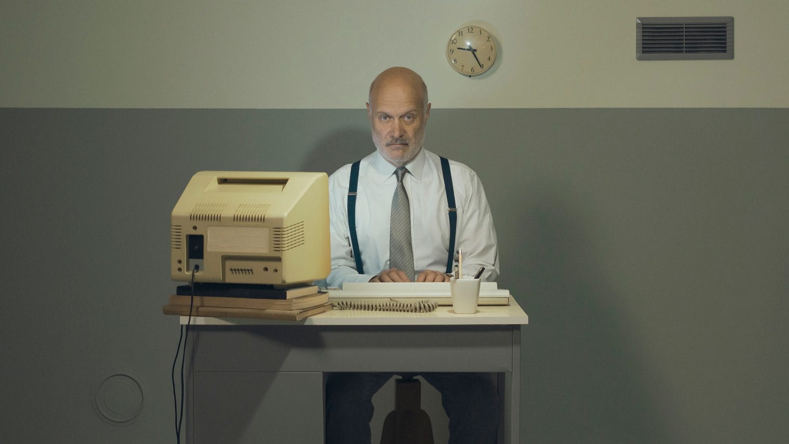 A businessman sits at an office desk that's way too small to get any work done.